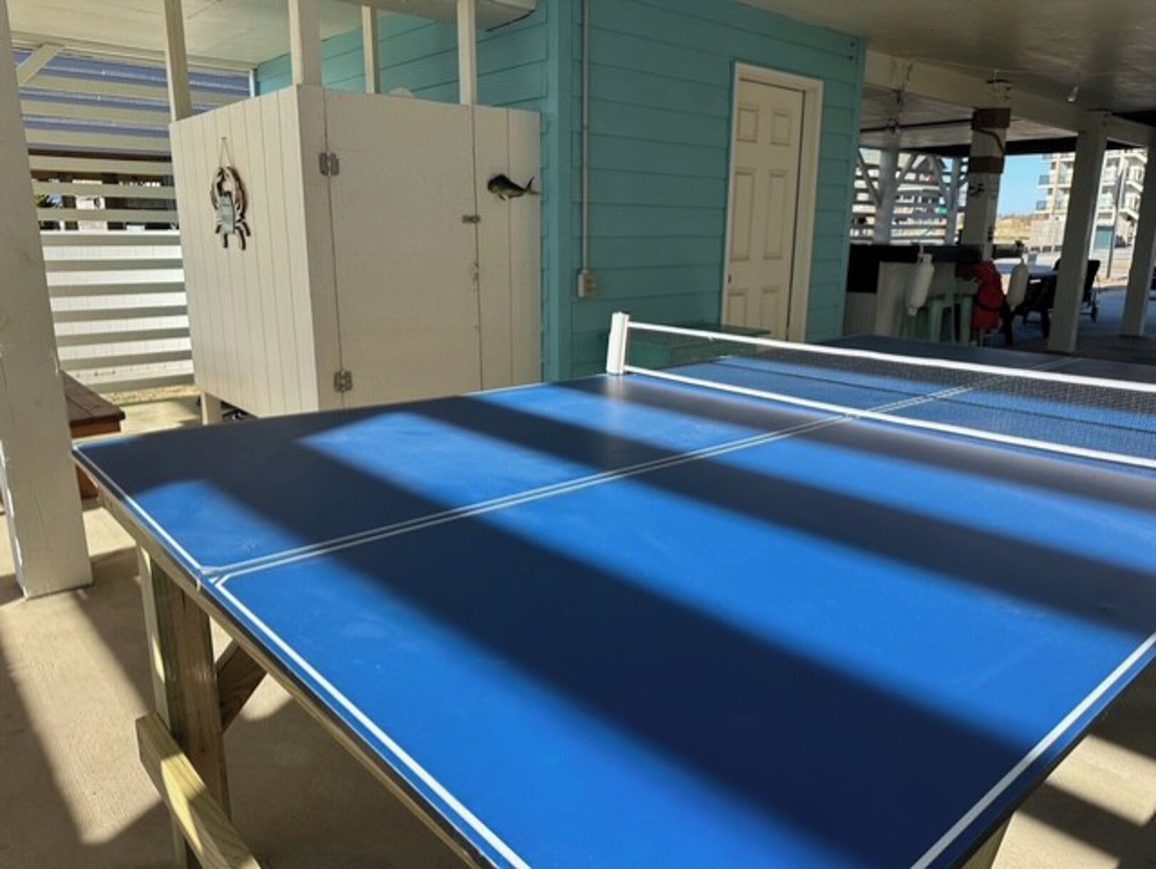 A blue ping pong table in a covered outdoor area with a white door and a small white building in the background.
