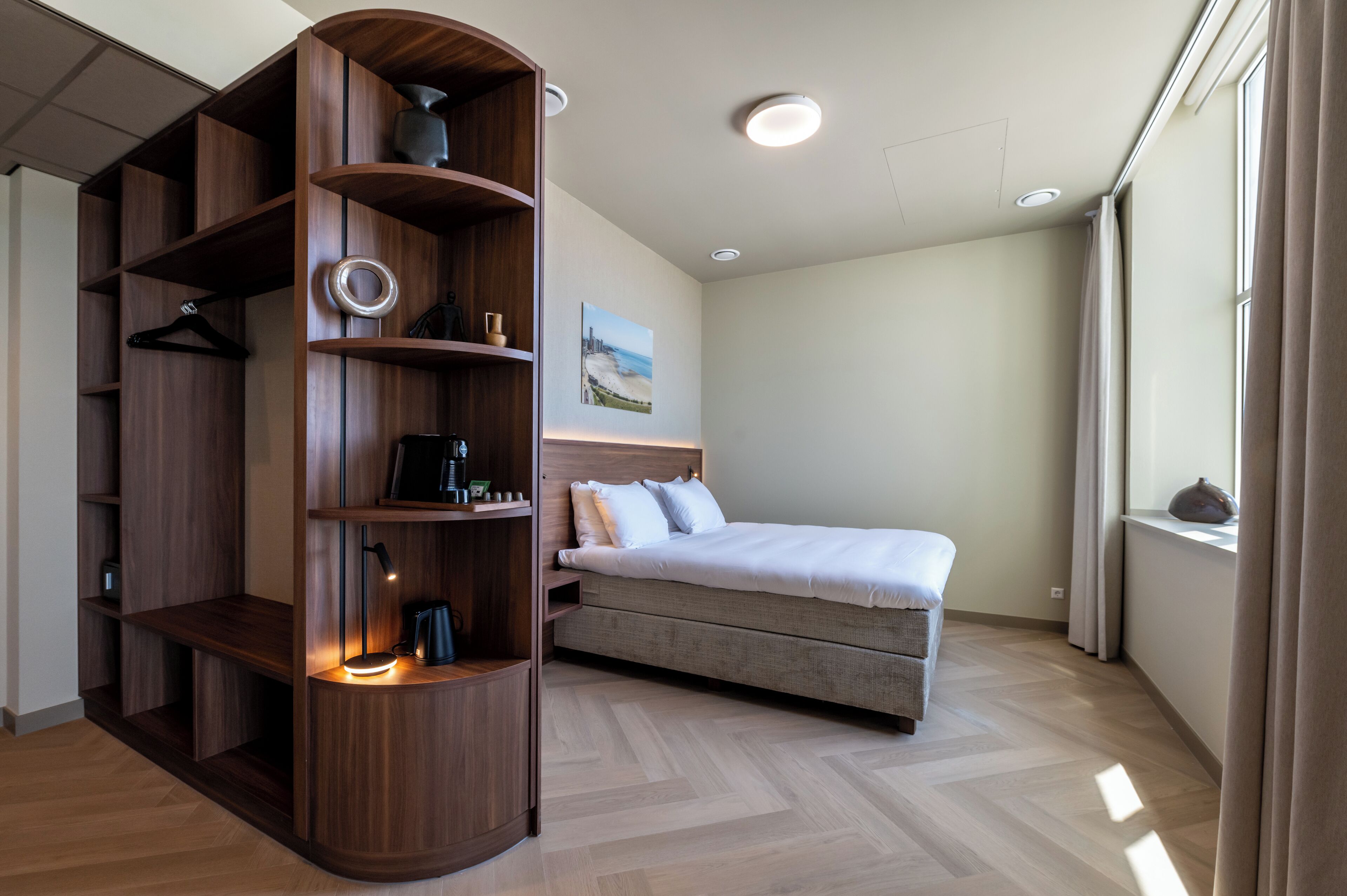 A bedroom with a bed, a wooden wardrobe, a painting on the wall, and a window with curtains.