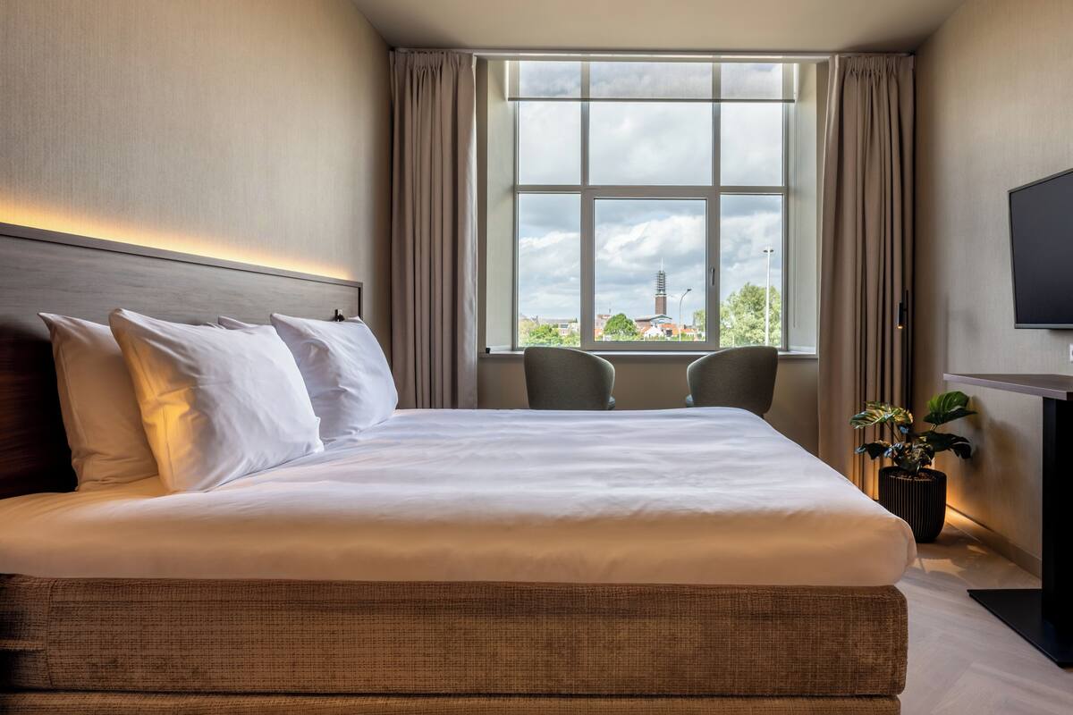 Ett hotellrum med en stor säng, två stolar, en TV och ett fönster med utsikt över en byggnad.
