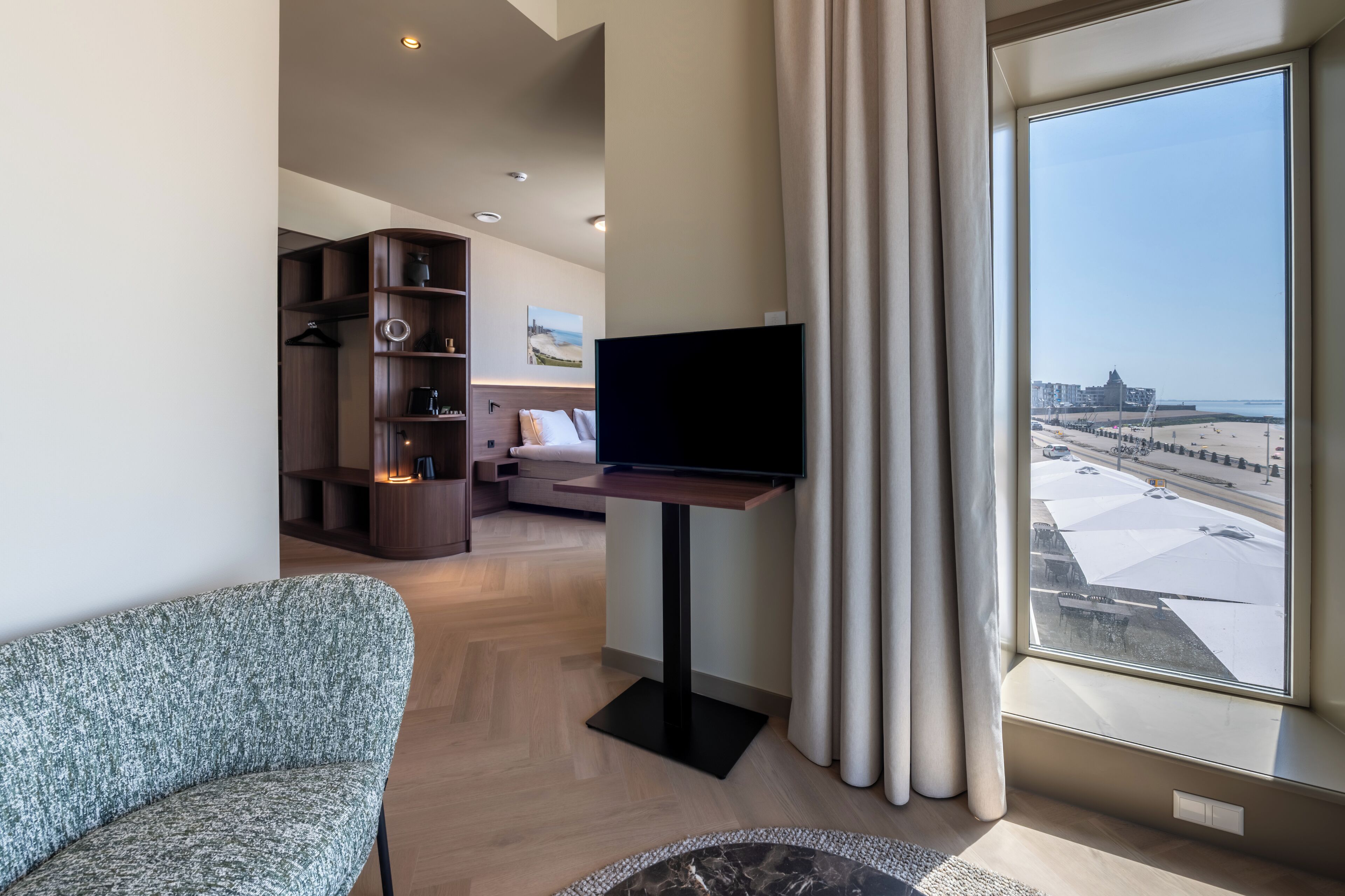 Een moderne hotelkamer met een groot raam, een flatscreen televisie, een bank, een bijzettafeltje en een ingebouwde boekenkast.