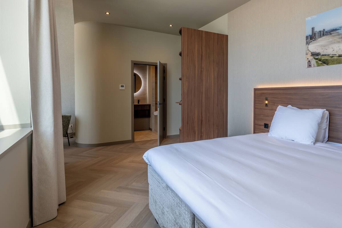 Ett hotellrum med en stor säng, en sänggavel i trä, ett badrum med en rund spegel och utsikt över stranden genom ett fönster.