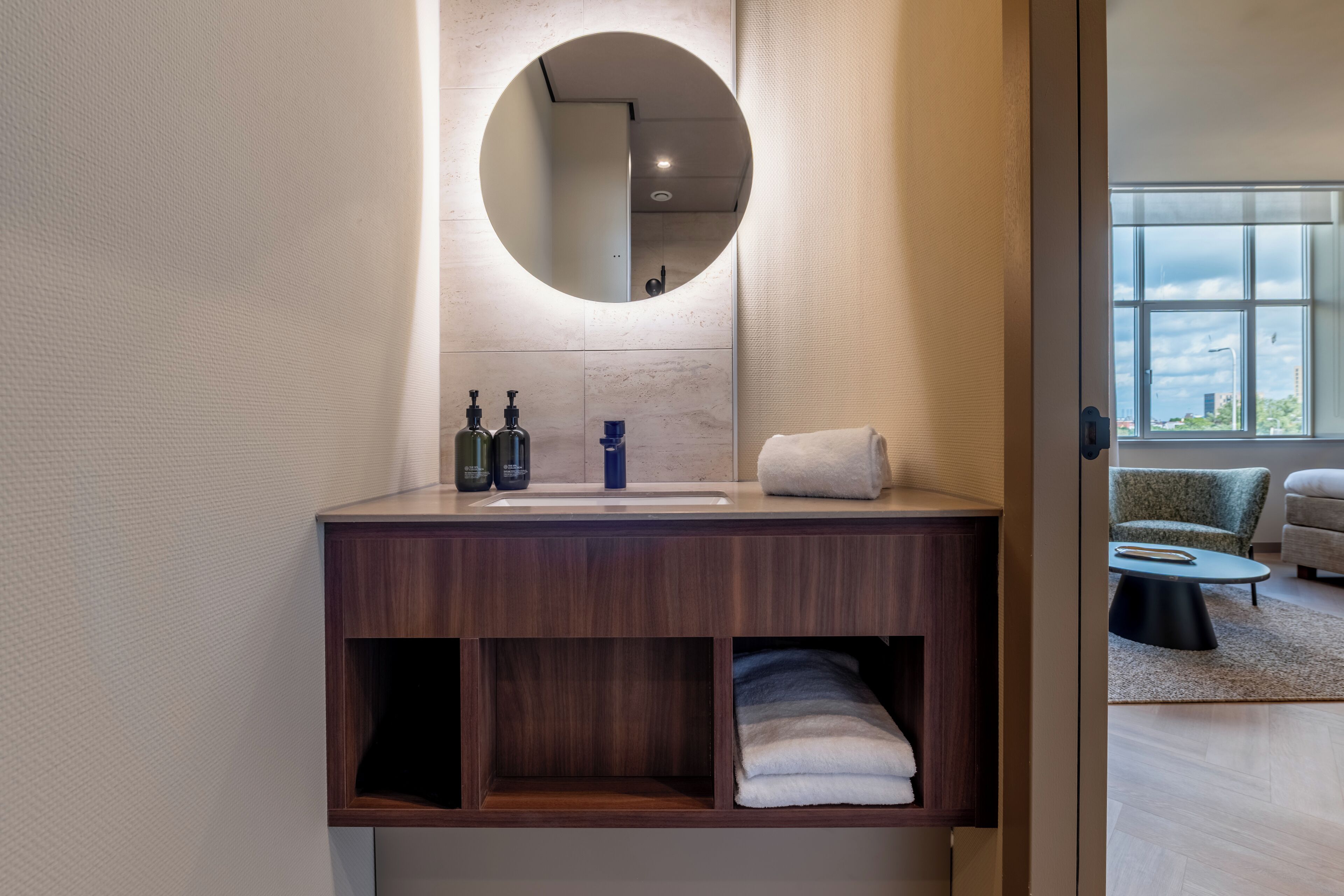 A modern bathroom with a wooden vanity, a round mirror, and a view of a living area through an open door.