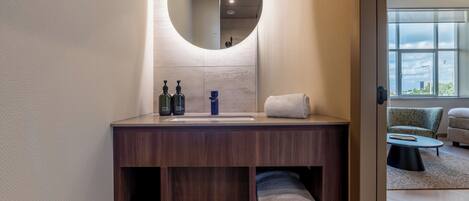 A modern bathroom with a wooden vanity, a round mirror, and a view of a living area through an open door.