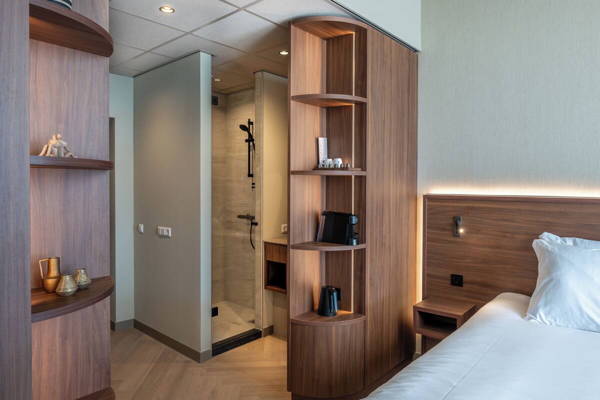 Ett modernt hotellrum med en garderob i trä, en rymlig klädkammare, en dusch och en snyggt bäddad säng.