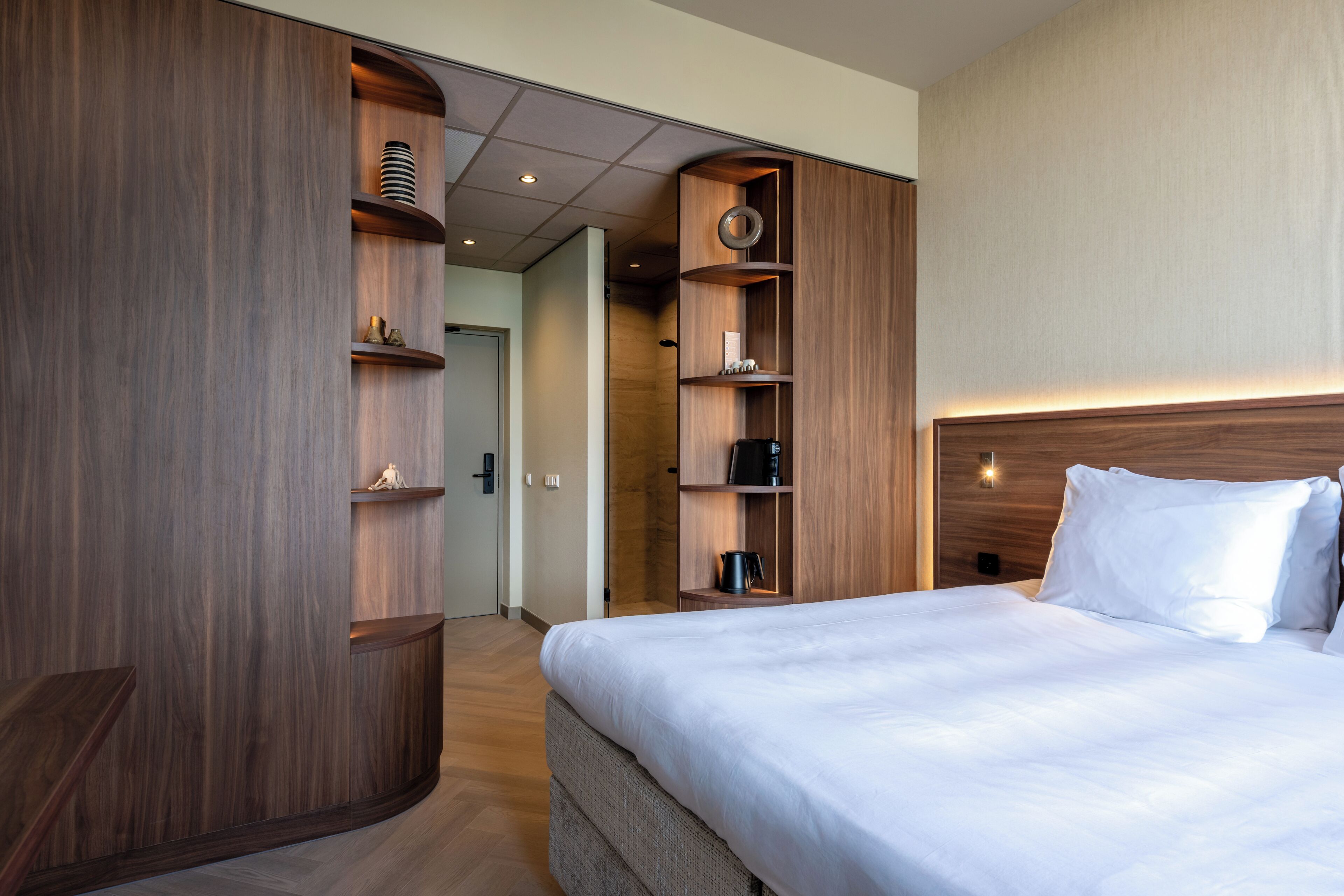 Une chambre d’hôtel avec un grand lit, une armoire en bois et des étagères intégrées où sont rangés divers objets.