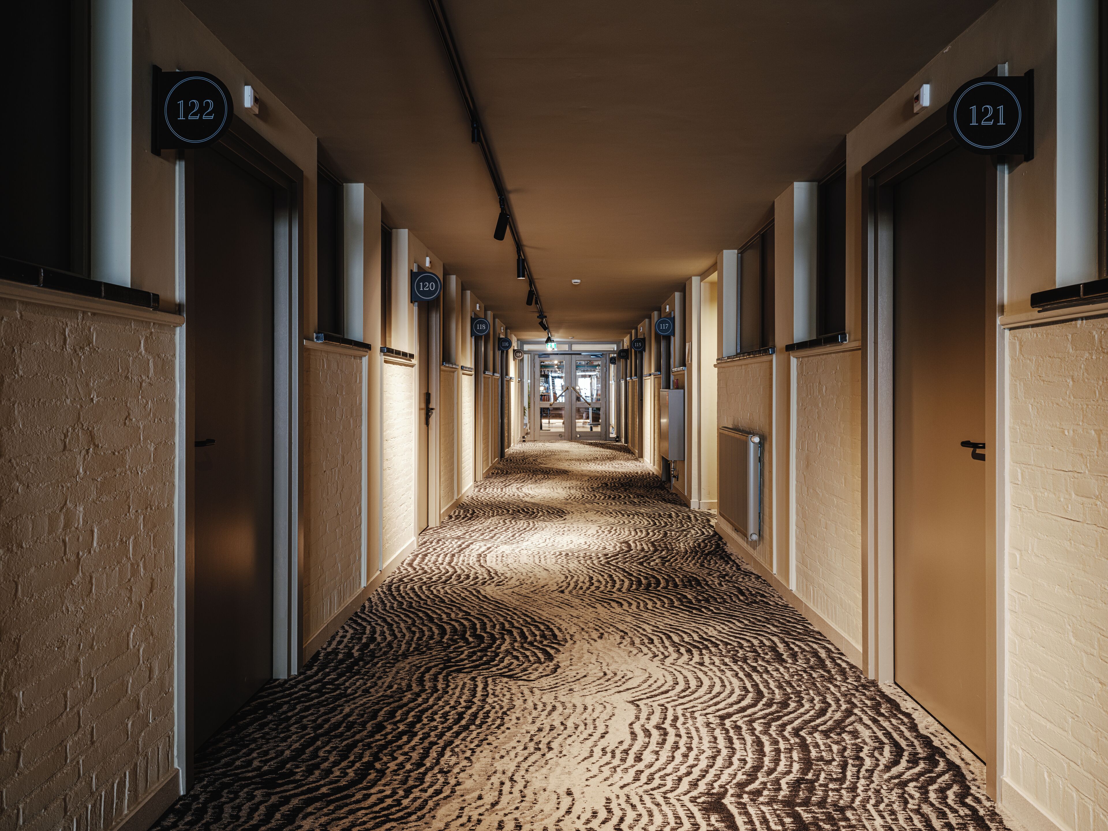 A hotel hallway with numbered doors, carpeted floors, and a brick wall on one side.