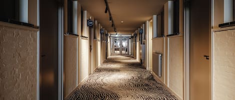 Un pasillo de hotel con puertas numeradas, piso alfombrado y una pared de ladrillo en un lateral.
