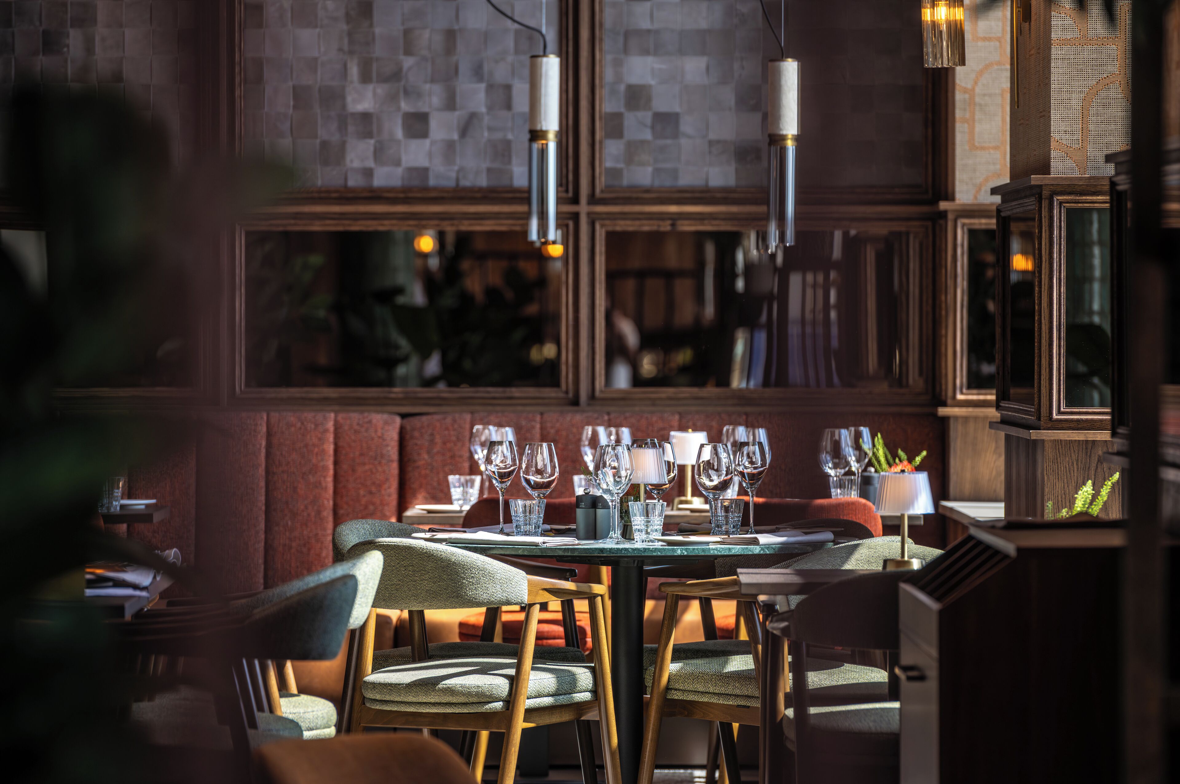 A dining area with a table set for four, wooden chairs, hanging pendant lights, and a view through a window with a patterned wall.