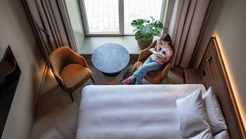 Una habitación de hotel con cama, una mesa redonda, una silla, una planta en maceta y una ventana con cortinas.