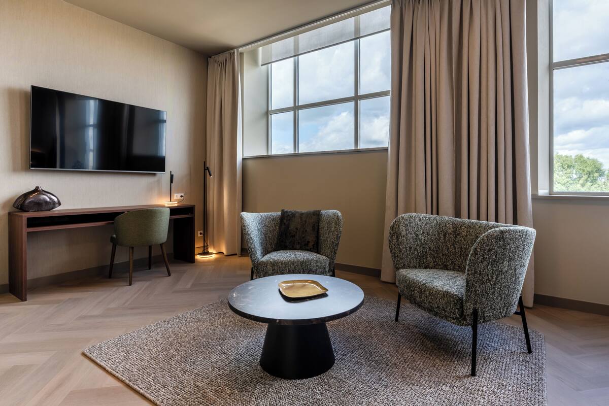 Ett modernt hotellrum med en platt-TV, ett skrivbord, två mönstrade fåtöljer och ett runt soffbord.