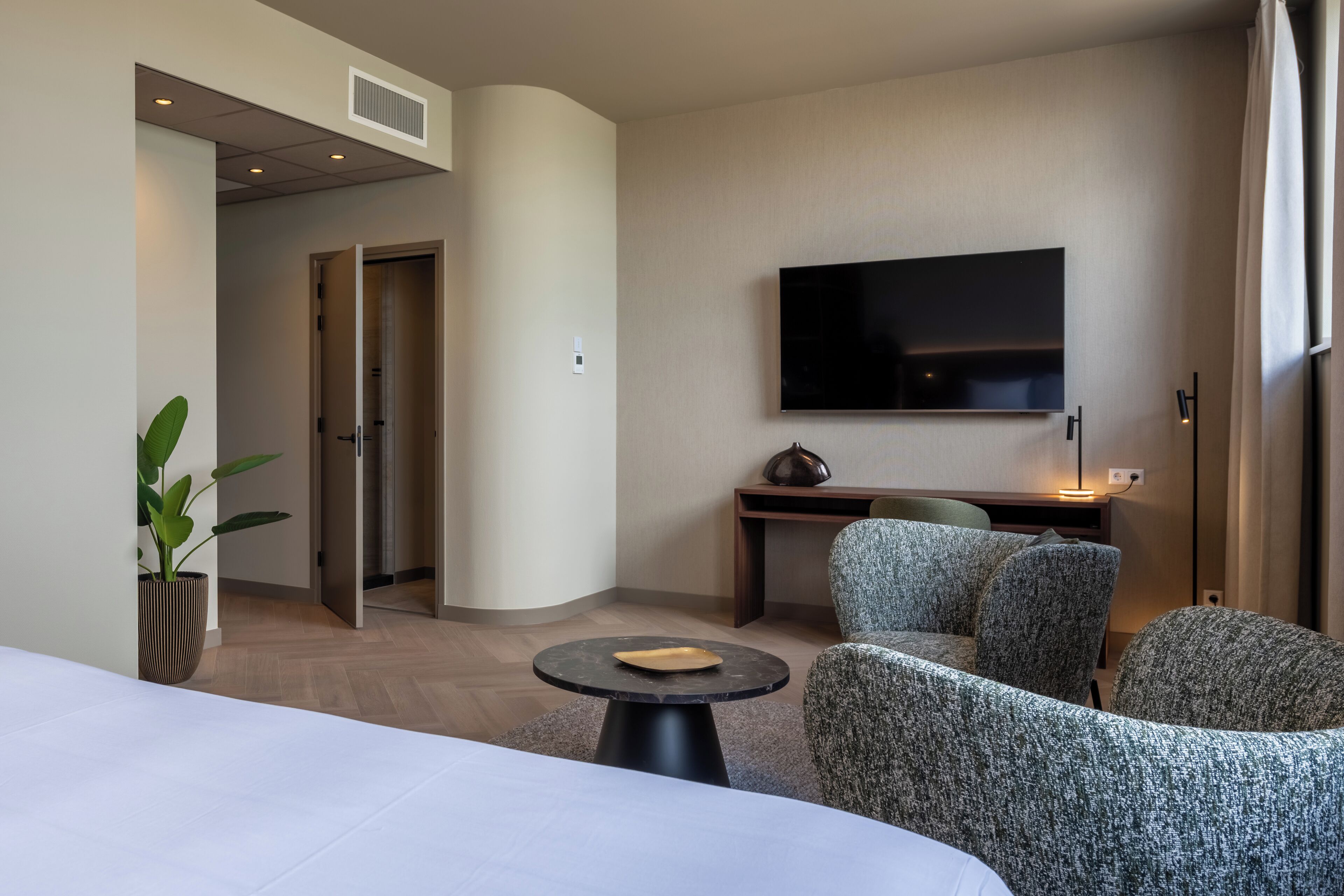 Una habitación de hotel moderna con una cama, una mesa redonda con un plato, dos sillones tapizados, un escritorio con una televisión de pantalla plana y una planta en maceta.