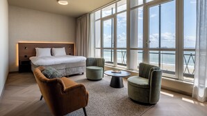 A modern hotel room with a large bed, two armchairs, a small coffee table, and a view of the ocean through large windows.