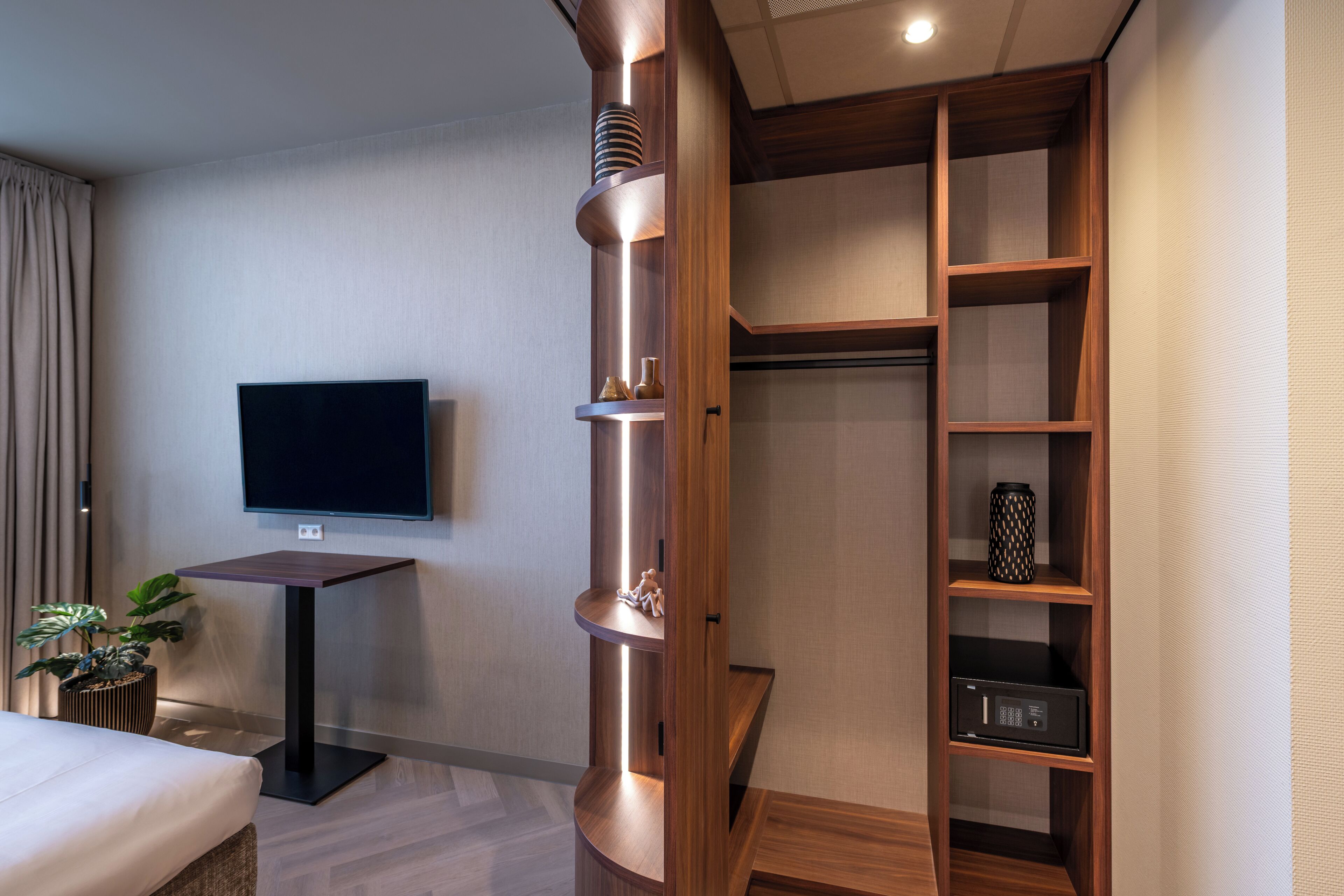 Una camera d'albergo moderna con una TV a schermo piatto fissata al muro, un mobiletto a mensole in legno con oggetti decorativi e un piccolo tavolino con una pianta in vaso.