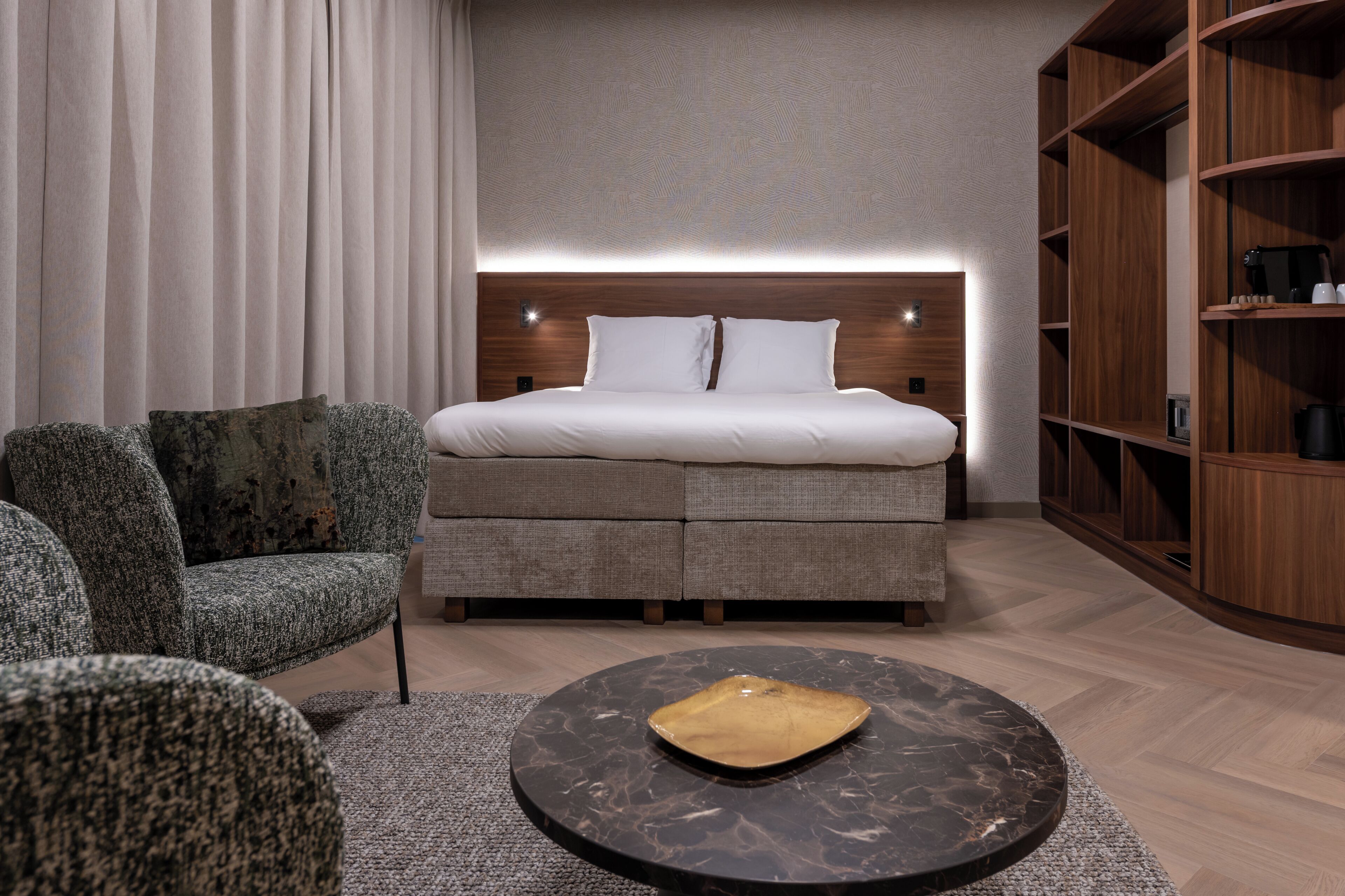 A modern hotel room with a bed, a round coffee table, a sofa, and a wooden bookshelf.