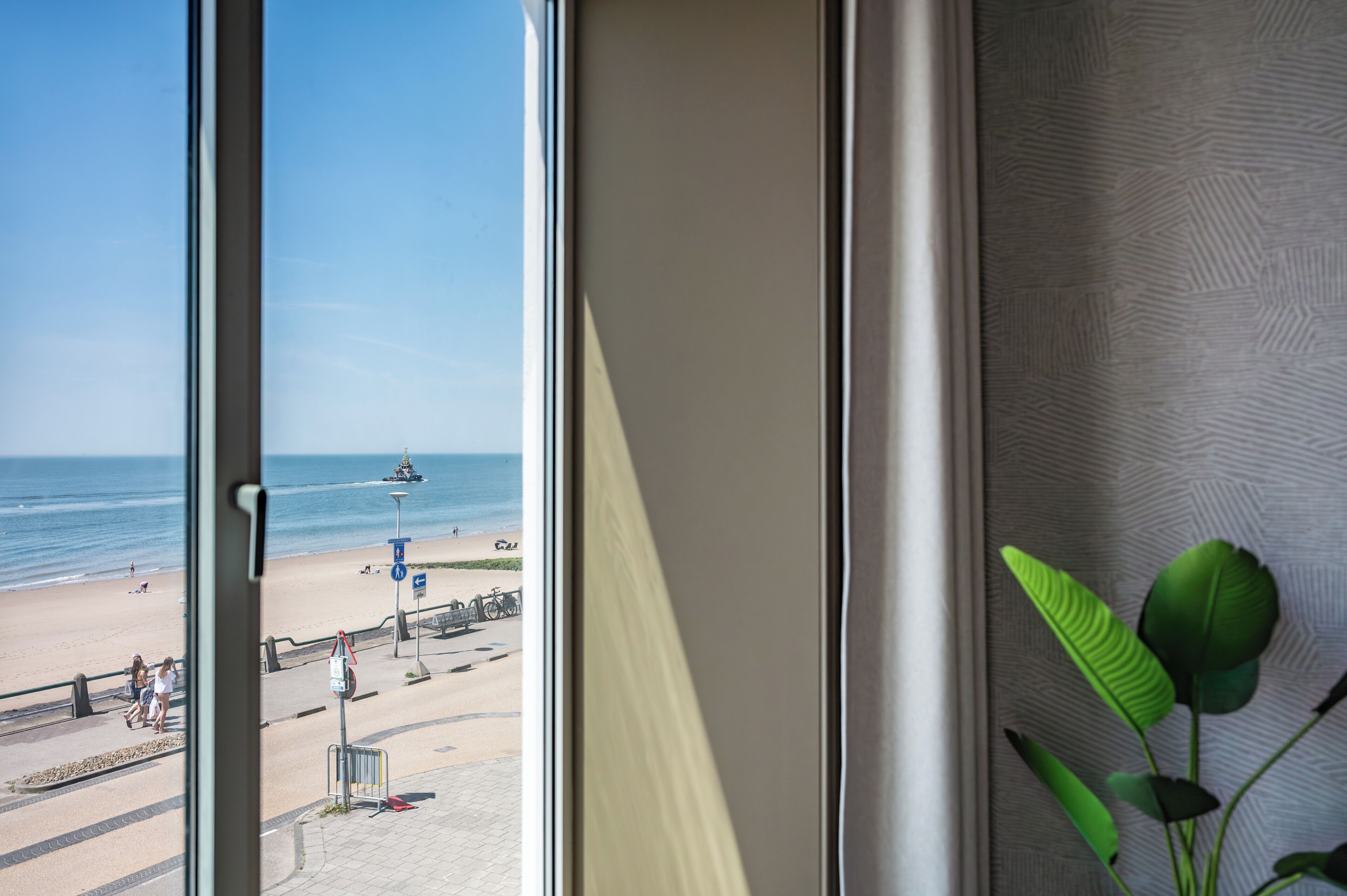 A view from a window overlooking a beach with a clear sky, a sandy shore, and a few people walking.