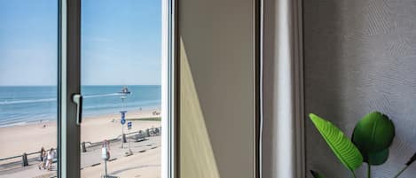 A view from a window overlooking a beach with a clear sky, a sandy shore, and a few people walking.