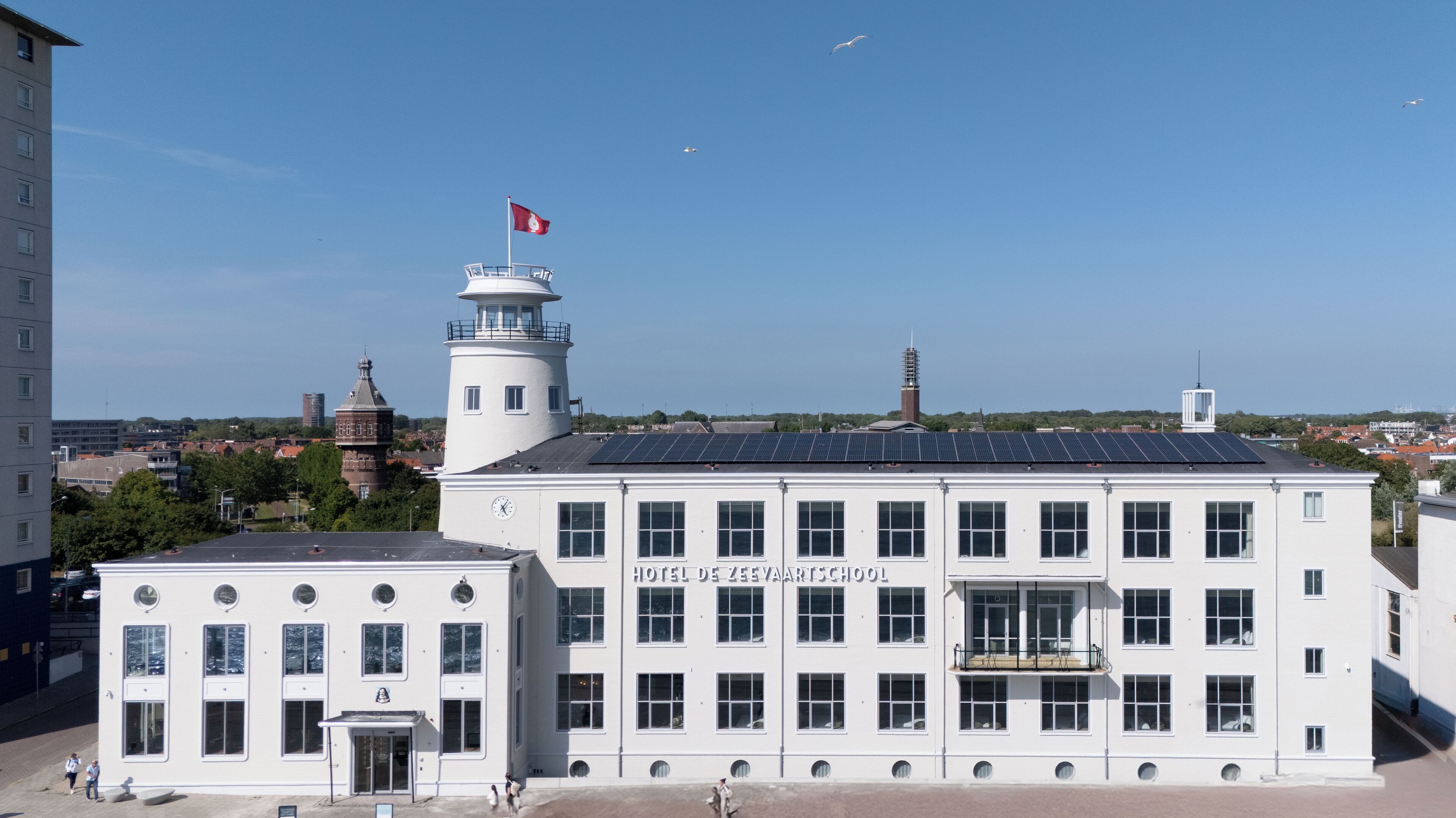 A white building with a clock tower and a sign reading "Hotel De Zeevaartschool".