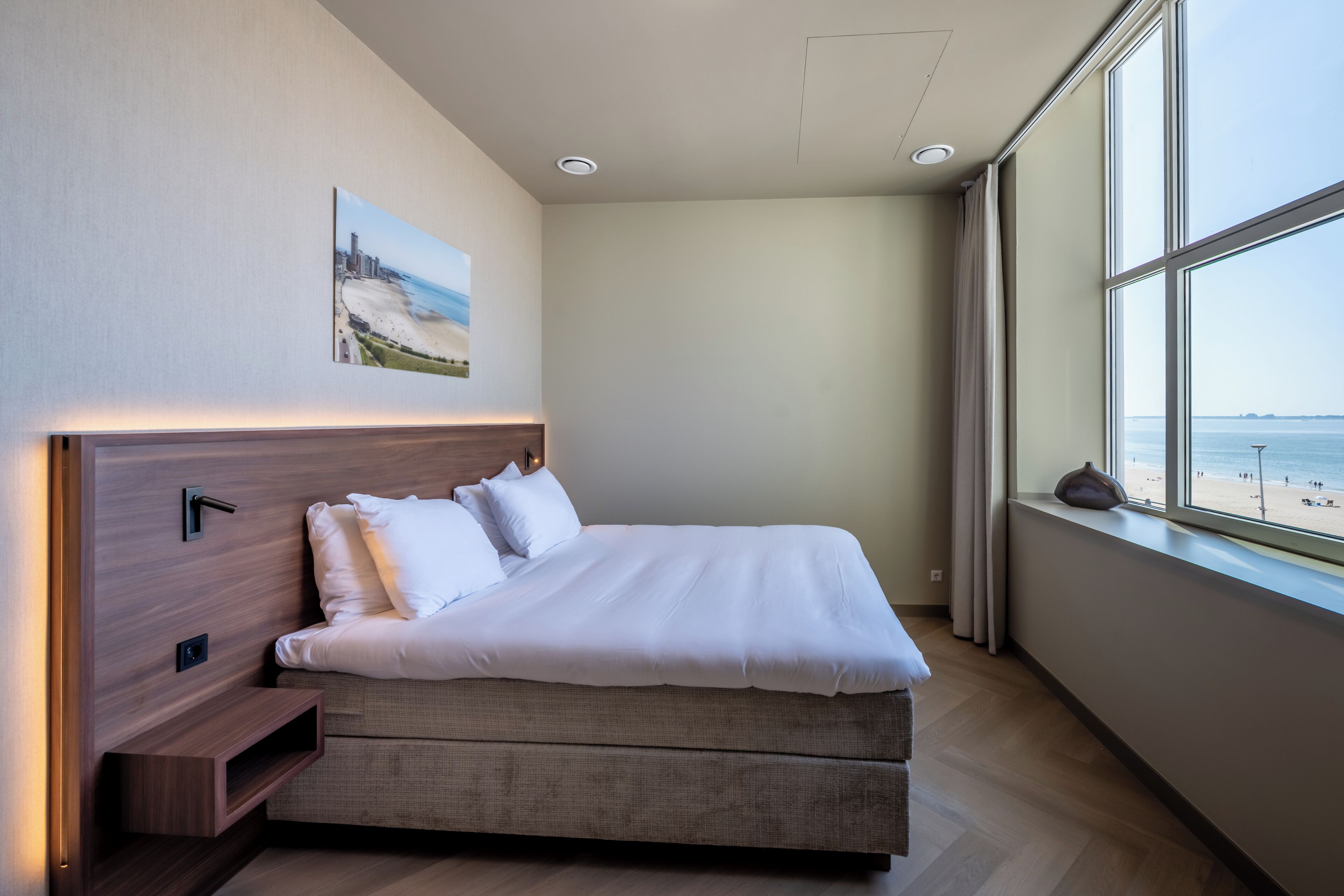 Ein modernes Hotelzimmer mit einem großen Bett, einem Nachttisch, einem Wandbild und einem Fenster mit Blick auf den Strand.