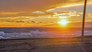 Een strandscène met de ondergaande zon boven de oceaan, golven die op het strand breken, en een paal op de voorgrond.