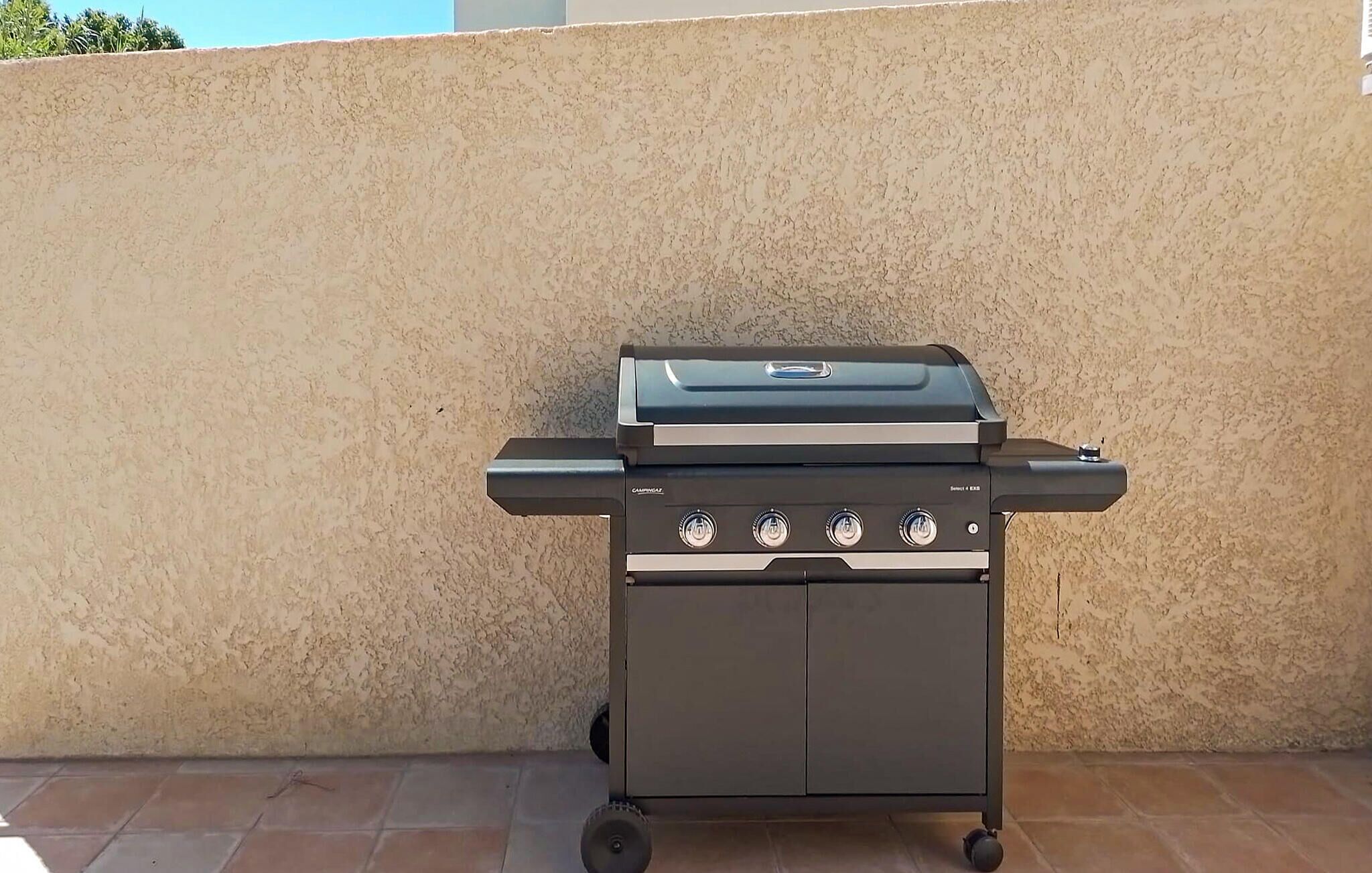 Una parrilla portátil con cuatro quemadores, una tapa y un estante lateral, ubicada sobre un patio con baldosas frente a una pared con textura.