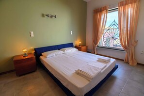 A neatly made bed with white linens, two bedside tables with lamps, a window with curtains, and a wall-mounted light fixture.