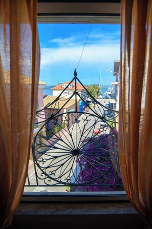 A window with a wrought iron gate, a view of a coastal town, and buildings with red-tiled roofs.