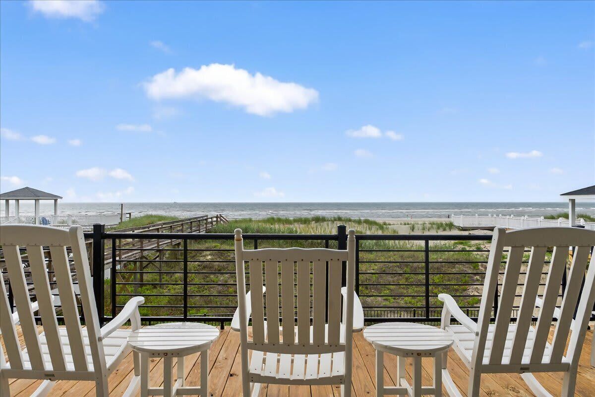 A deck with three white chairs and a table, overlooking a beach with a wooden walkway and grassy dunes.