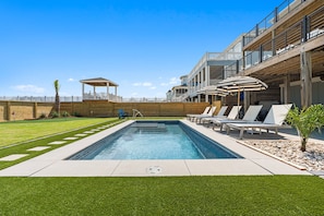 A swimming pool with lounge chairs and umbrellas, a wooden deck, and a covered seating area. - Shell Yeah- Ocean Isle Beach (Ocean Isle Beach)