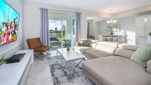 A modern living room with a large TV, a brown leather chair, a beige sofa, and a glass coffee table. - Modern Condo Near Disney (Kissimmee)