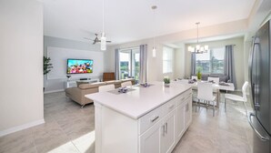 A modern kitchen with a central island, a dining area with a white table and chairs, and a living area with a sofa and TV. - Modern Condo Near Disney (Kissimmee)