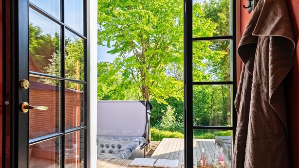A glass door leading to a wooden deck with a hot tub, a brick wall, and a tree in the background.