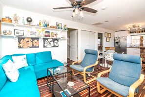 A living room with a blue sofa, a glass coffee table, and two blue armchairs. There is a ceiling fan, a kitchen area with a refrigerator, and a dining table with chairs. - Hibiscus Hut - 2 BR Cottage with Panoramic Ocean Views! Dog Friendly (North Topsail Beach)