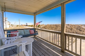 Eine Holzterrasse mit Schaukel und Tisch, mit Blick auf einen Strand mit Dünen und einem weißen Gebäude im Hintergrund.