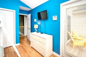 A room with a blue wall, a white dresser, a television mounted on the wall, and a window with blinds.