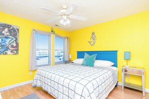Ein Schlafzimmer mit einem Bett, einem Deckenventilator, einem Fenster mit Jalousien, einem Wandbild und einem Nachttisch.
