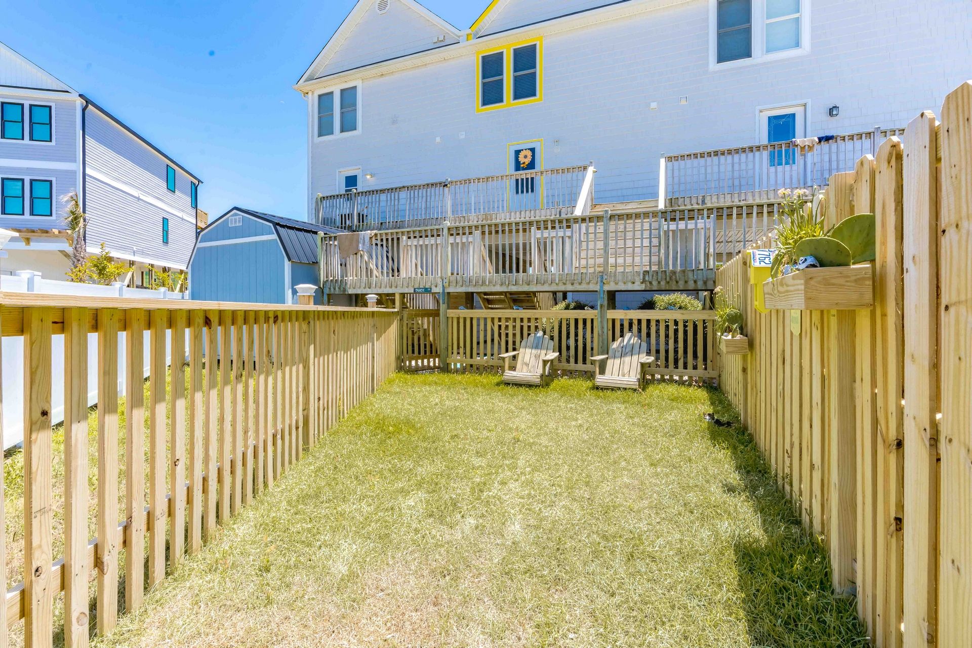 A fenced backyard with a grassy lawn, a wooden deck, and two Adirondack chairs.