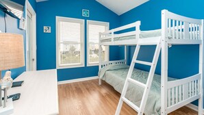 A room with two bunk beds, a desk with a lamp, and two windows with blinds.