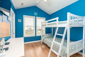 A room with two bunk beds, a desk with a lamp, and two windows with blinds.