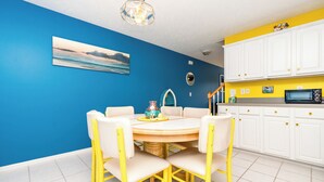 A dining area with a round table and chairs, a kitchen with white cabinets and a yellow countertop, and a blue wall with a painting.