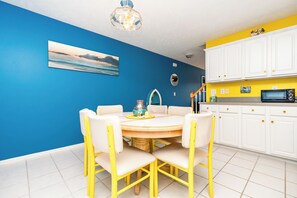 A dining area with a round table and chairs, a kitchen with white cabinets and a yellow countertop, and a blue wall with a painting.