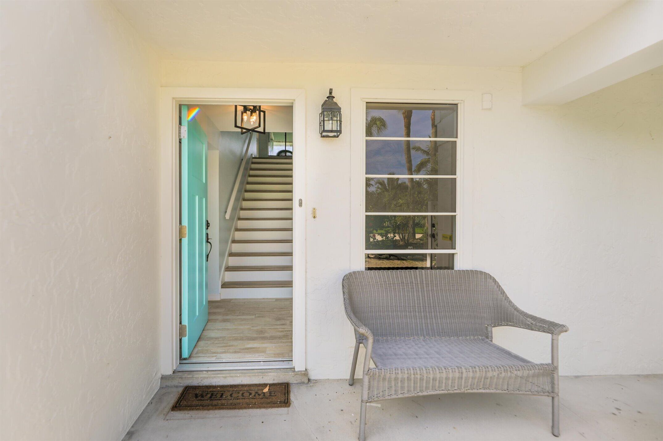 A covered porch with a wicker chair, a staircase leading to a door, and a window with a view of palm trees.
