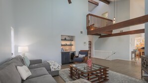Ein geräumiges Wohnzimmer mit grauem Sofa, einem Glastisch und einem Holzstuhl. Eine Treppe führt auf eine obere Ebene.
