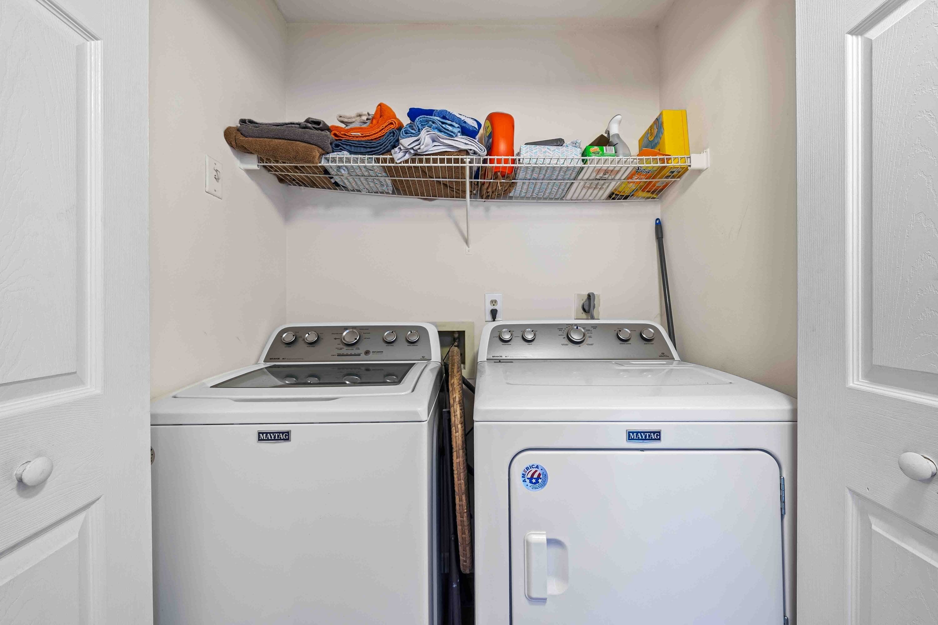 A laundry room with two Maytag washers and dryers, a metal shelf above holding folded clothes and cleaning supplies.