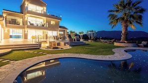 Une maison moderne à deux étages, dotée d’une piscine, d’un espace salon extérieur et d’un palmier.