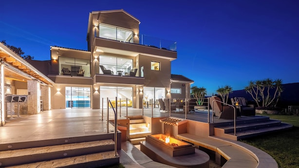 Une maison moderne à deux étages, dotée d’une vaste terrasse extérieure, d’un foyer et d’une entrée bien éclairée.
