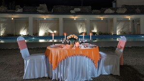 An outdoor dining setup with a table set for two, candles, and a view of a building with a pool in the background. - Vanashraya corbett (Kaladhungi)