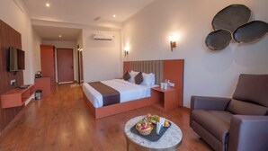 A modern hotel room with a large bed, a seating area with a brown leather chair, a small table with fruit and snacks, and a TV mounted on the wall. - Vanashraya corbett (Kaladhungi)