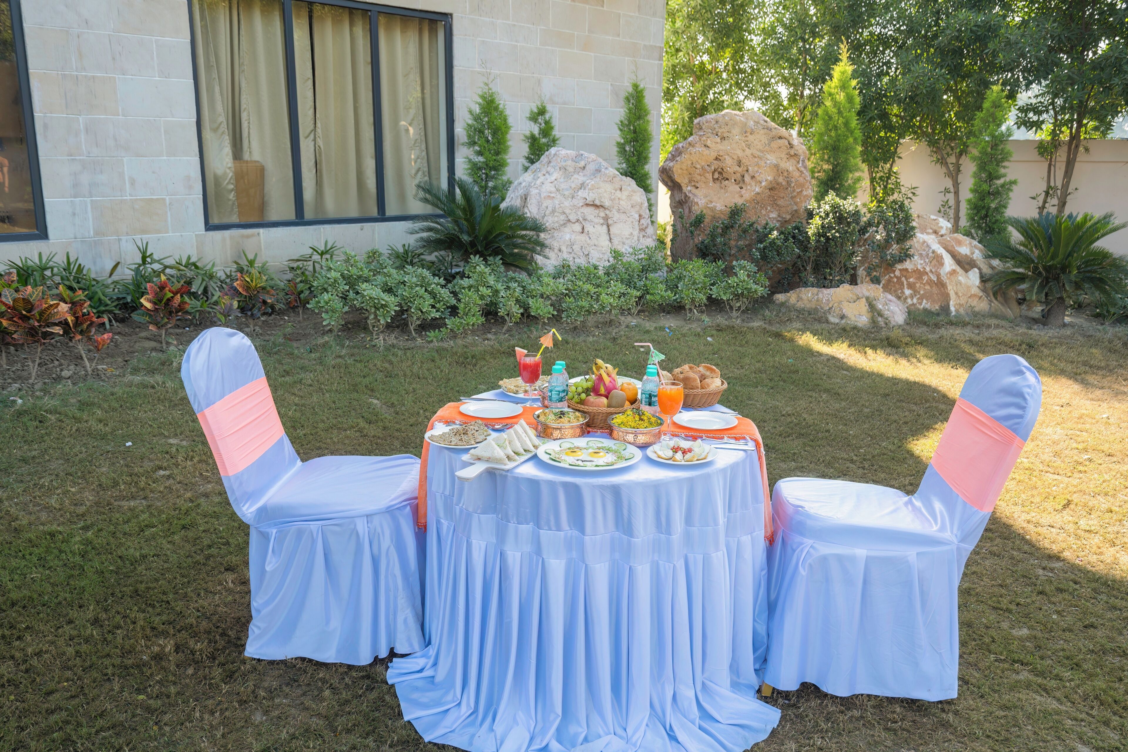 An outdoor dining area with a table set for a meal, surrounded by chairs with pink sashes, and a stone building in the background.
