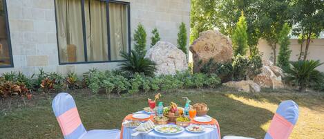 An outdoor dining area with a table set for a meal, surrounded by chairs with pink sashes, and a stone building in the background.