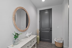 A bathroom with a white vanity, a round mirror with a wooden frame, a white decorative fish, a vase with green plants, and a grey door.